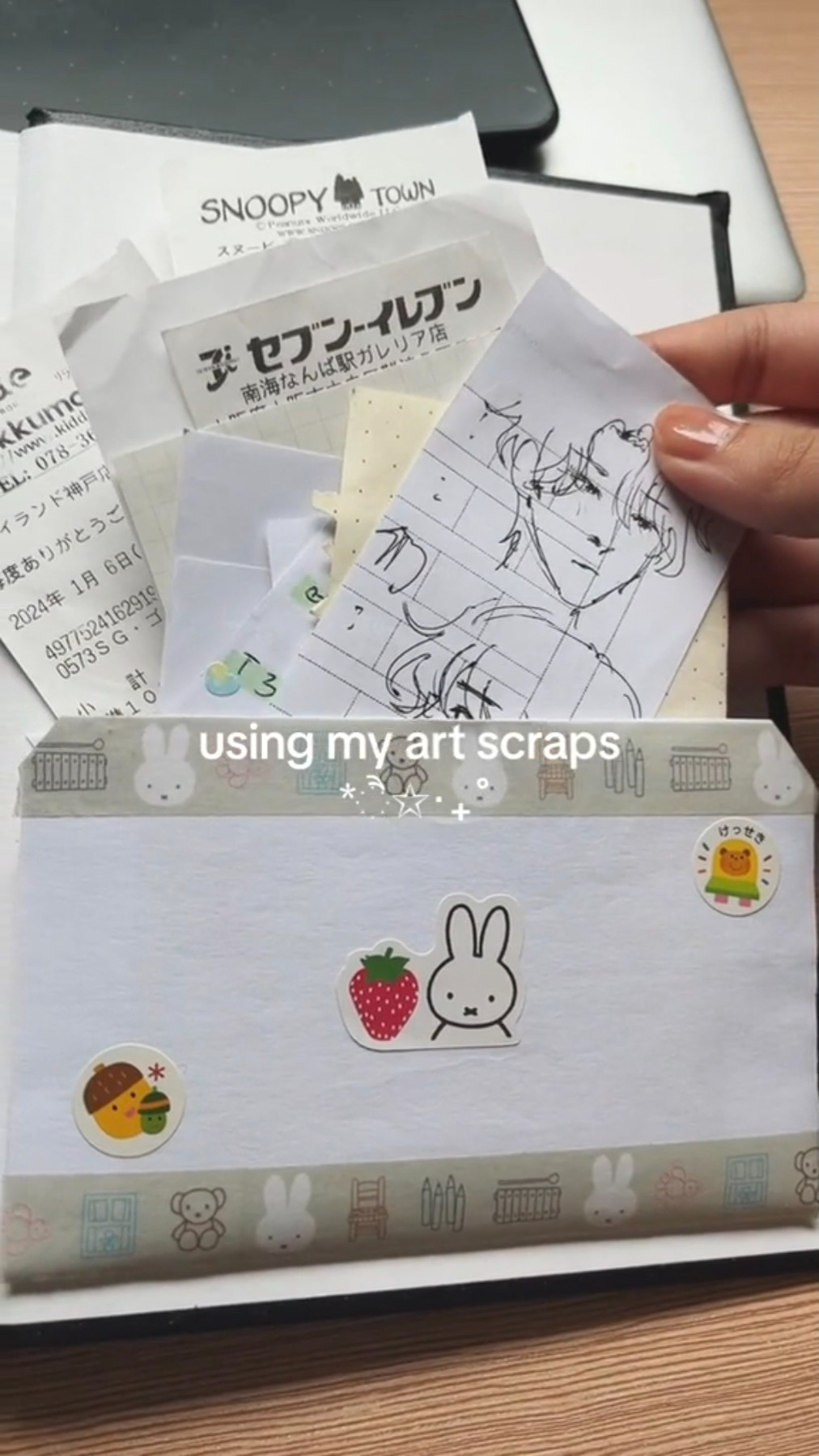 a hand holding a collection of papers with doodles, stickers and other art supplies being added to a planner or journal. the hand is holding a piece of paper with a doodle of a character with short hair. it also has a small notebook, a receipt and a sticker from a convenience store.  the text "using my art scraps" is visible below the items.
