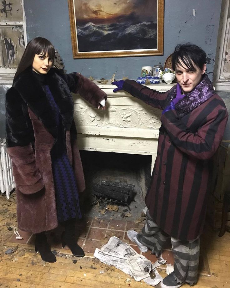 the image shows two people, a woman and a man, standing in front of a fireplace. the woman on the left is wearing a large, twotone coat with dark brown fur trim, a dark dress, and black boots. she has dark hair and bangs. the man on the right is wearing a dark pinstripe coat, striped pants, black shoes, and a purple scarf and gloves. the fireplace is white and ornate, with a painting of a seascape hanging above it. the floor is littered with debris. the setting appears to be a somewhat dilapidated room. it is likely a scene from the tv series "gotham", with the actors playing morena baccarin and robin lord taylor.