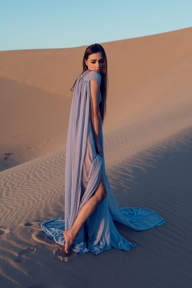 a woman in a long, flowing periwinkle dress stands on a sand dune in the desert. the dress is sheer and billows in the wind, revealing the contours of her body. she is looking down, her long brown hair cascading over her shoulders. the sand is a soft, golden brown, and the sky is a clear, pale blue. the sun is setting, casting a warm, golden glow over the scene.
