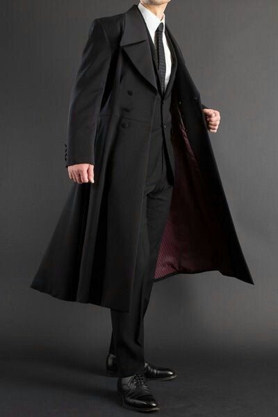 a man wearing a long black coat, a formal suit, and a tie is standing in front of a dark grey background. he is also wearing black shoes.