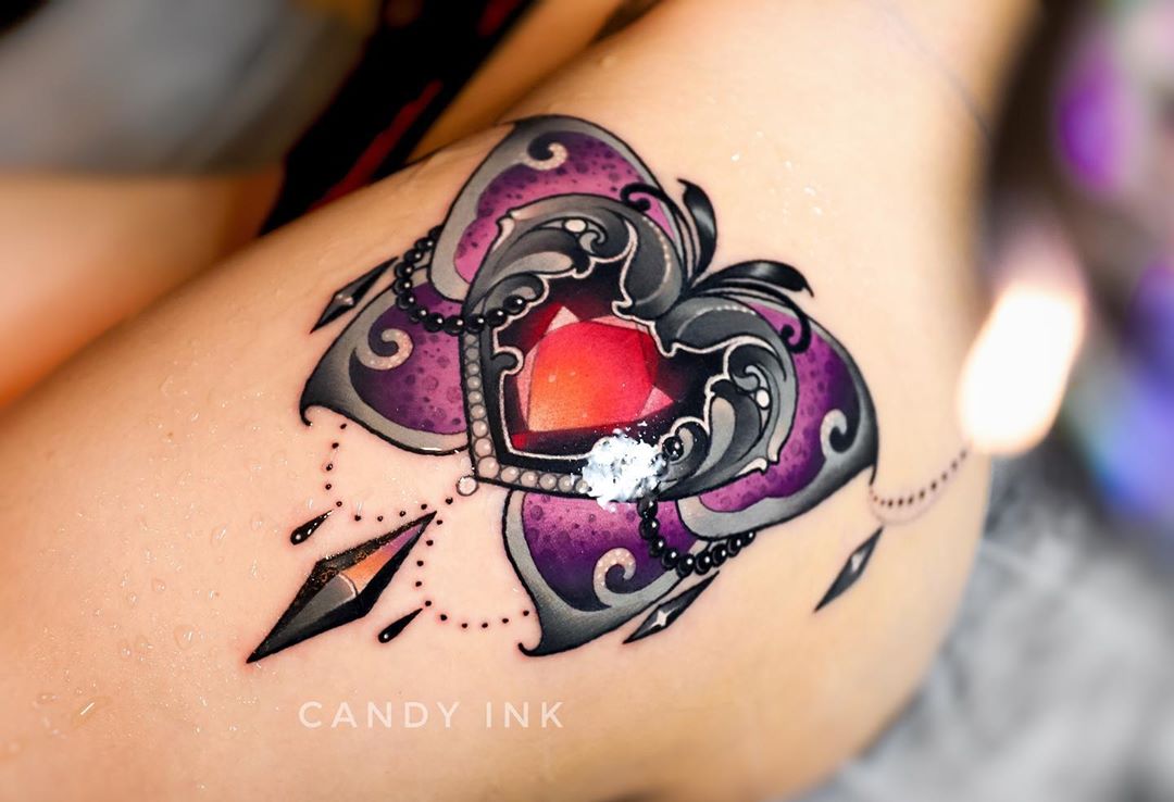a closeup photograph showcasing a neotraditional tattoo of a heartshaped jewel nestled within the framework of a butterfly. the artwork, executed in a vibrant palette of purple, silver, and black, is situated on the upper thigh of a woman.