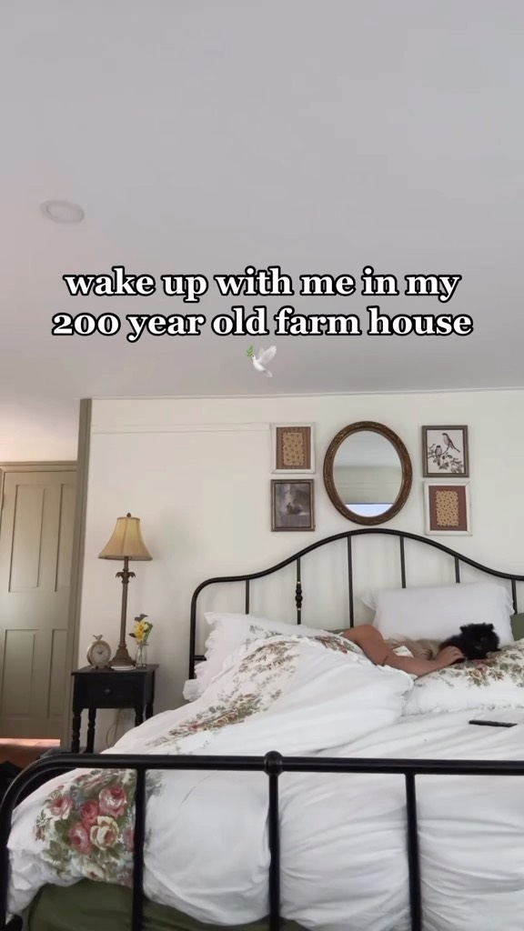 a bedroom scene in a farmhouse. a person is lying in a bed with white floral bedding. behind the bed are several pieces of wall decor, including framed pictures and an oval mirror. to the left of the bed, there is a nightstand with a lamp and a clock. the bedroom is decorated in a vintage or rustic style, characteristic of a farmhouse interior.