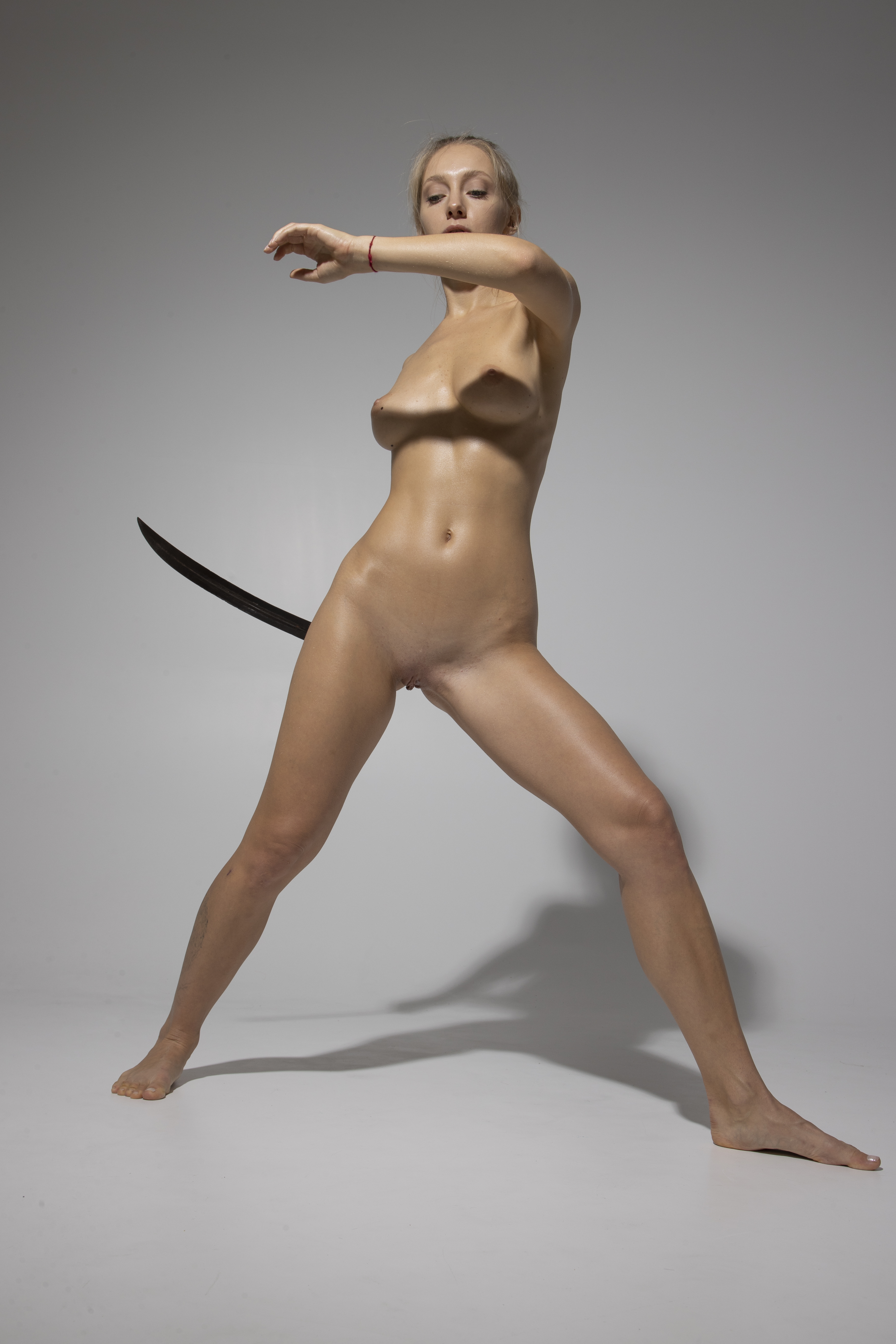 a full shot of a nude woman standing in a wide stance with a sword behind her back. she has blonde hair pulled back and is facing forward with her arm raised. the background is a plain grey, and the lighting is soft. she has a thin red bracelet on her wrist. the sword is black and appears to be a katana style weapon. the woman has a strong and powerful pose, exuding confidence.