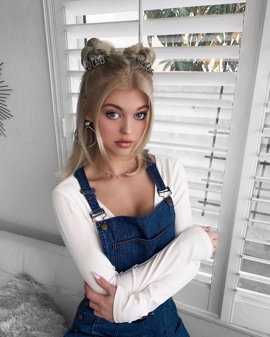 a young woman with blonde hair styled in two buns, accessorized with hair clips, posing in front of a window with white shutters. she's wearing denim overalls over a white longsleeve shirt, a necklace, and hoop earrings.