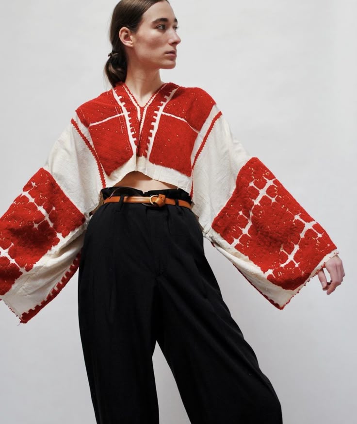 a woman is modeling a white crop top with red embroidery and black wideleg pants. she is wearing a brown leather belt. she is standing in front of a white background.