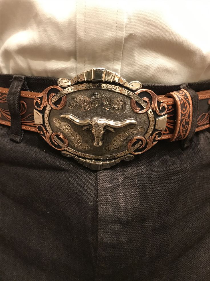 loseup view of a person wearing a brown leather belt with a large, ornate silver and copper belt buckle featuring a longhorn steer in the center. the buckle is highly detailed with engraved designs. the belt is worn over darkcolored jeans and a white shirt.  the focus is sharply on the buckle.