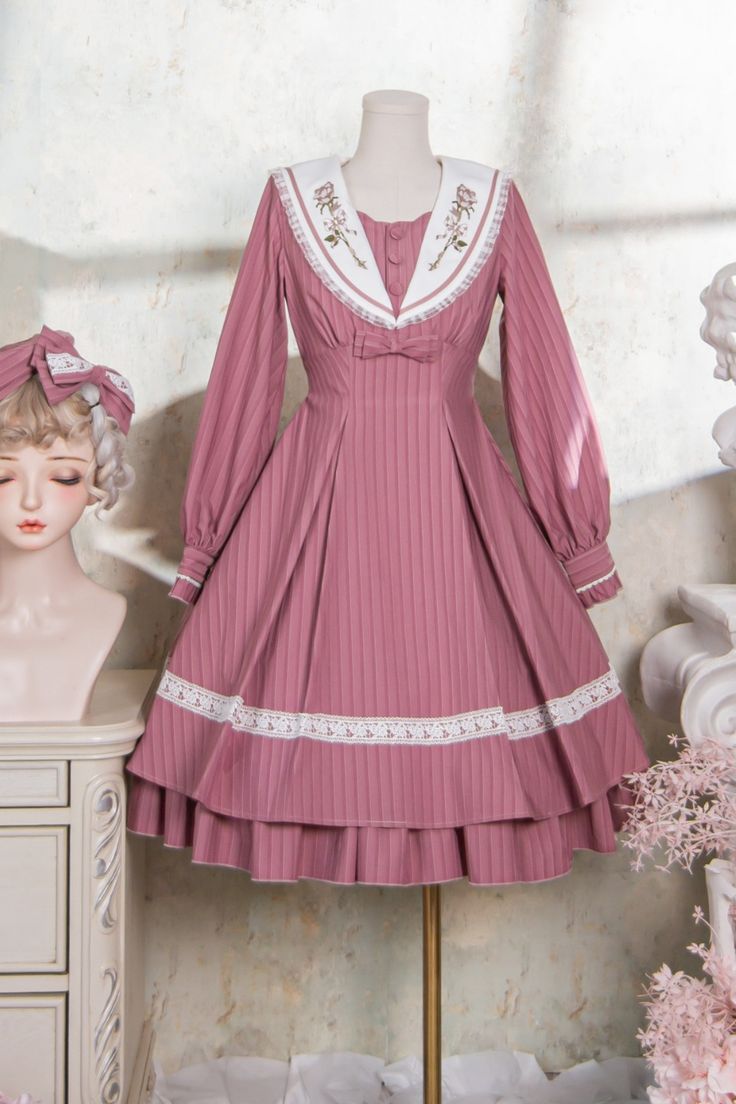 a pink lolita fashion dress with a sailor collar is displayed on a mannequin. the dress is photographed against a white wall with decorative molding. there are pink flowers to the right and a bust of a blonde woman wearing a matching headband to the left.  the dress has long sleeves, a square neckline, and a flared skirt that falls to the knee. the fabric is a pink and white striped pattern, and the dress is decorated with white lace and embroidery. the sailor collar is white with pink floral embroidery. the overall effect is sweet and feminine.