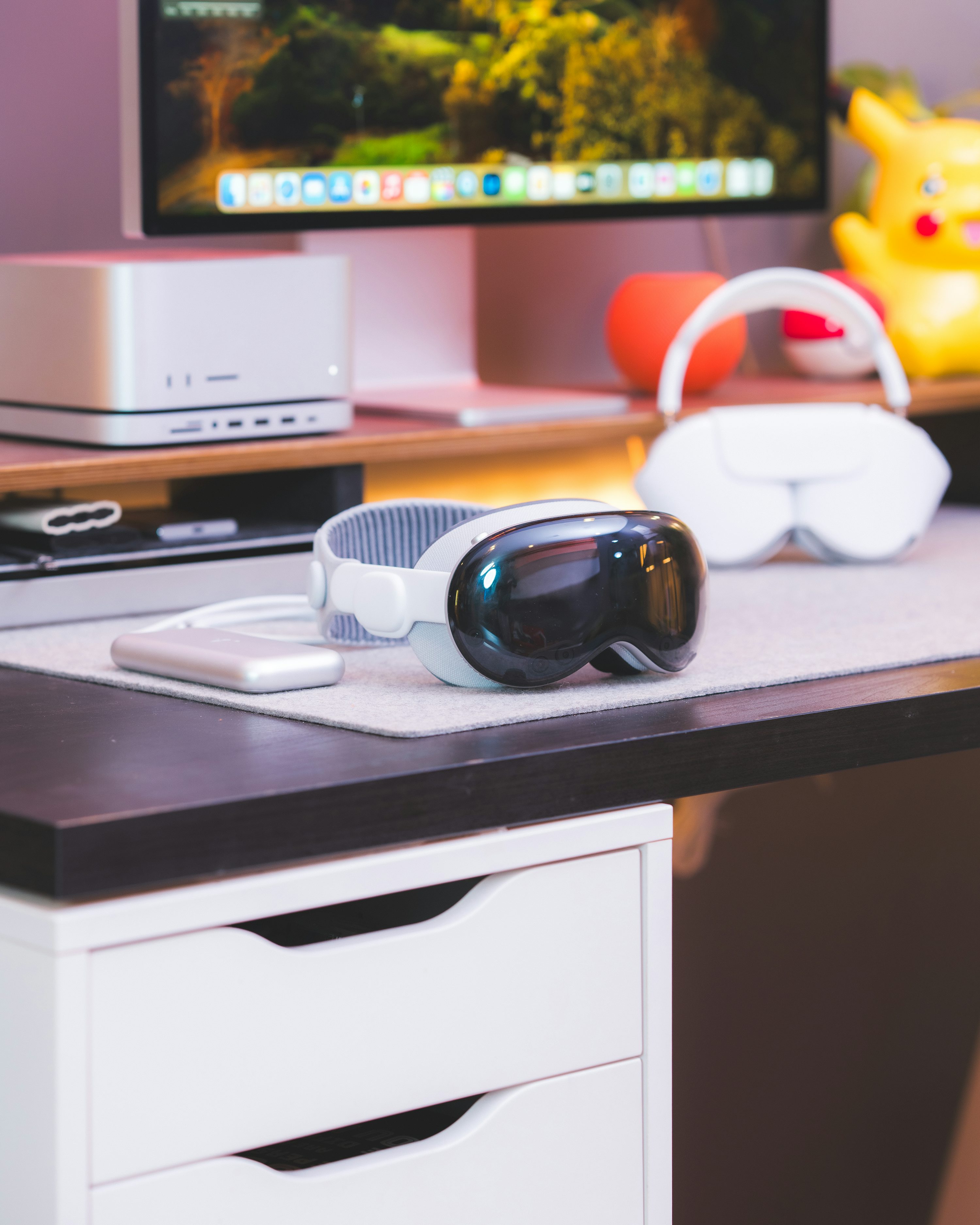the image shows a desk with a black top and white drawers. on the desk is a desk mat with an apple vision pro, an external hard drive, and apple airpods max. in the background are a mac studio, a computer screen, a pikachu figure, an orange decoration, and a poke ball decoration.