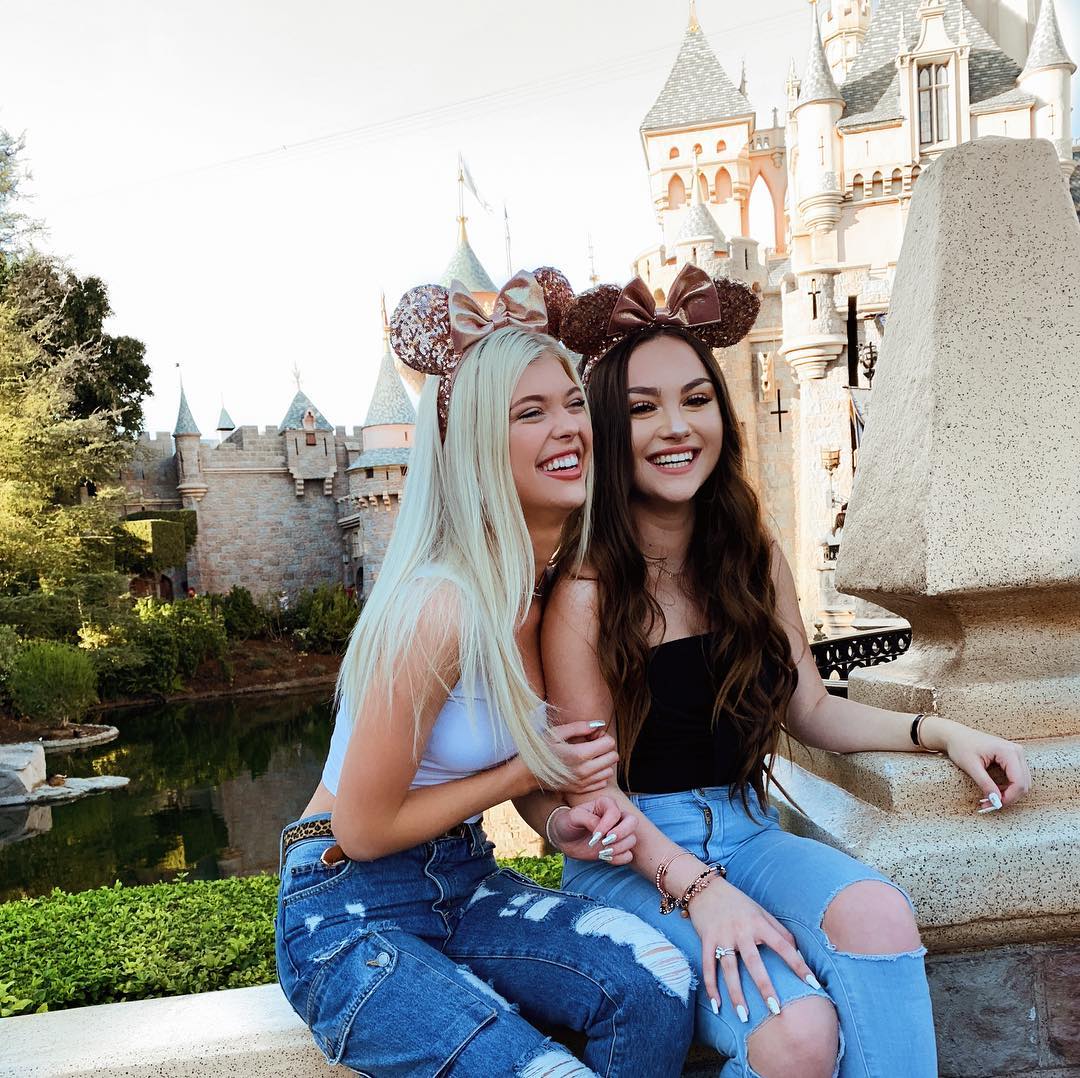 two young women wearing disney themed mouse ears are sitting in front of sleeping beauty castle at disneyland. they are smiling and appear to be enjoying their vacation. the woman on the left has blonde hair, ripped jeans and a white tank top. the woman on the right has brown hair, ripped jeans and a black tank top.