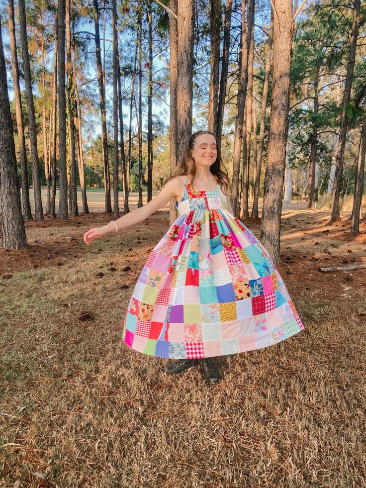 a full shot of a young woman wearing a patchwork dress, standing among tall trees in a forest. the woman is smiling with her eyes closed and her arms outstretched to the sides. she has long brunette hair and is wearing a simple bracelet. the dress is a colorful patchwork design, made of many small squares of different fabrics. the forest is filled with tall, thin trees and the ground is covered in fallen leaves and pine needles. the sunlight filters through the trees, creating a warm and golden light.