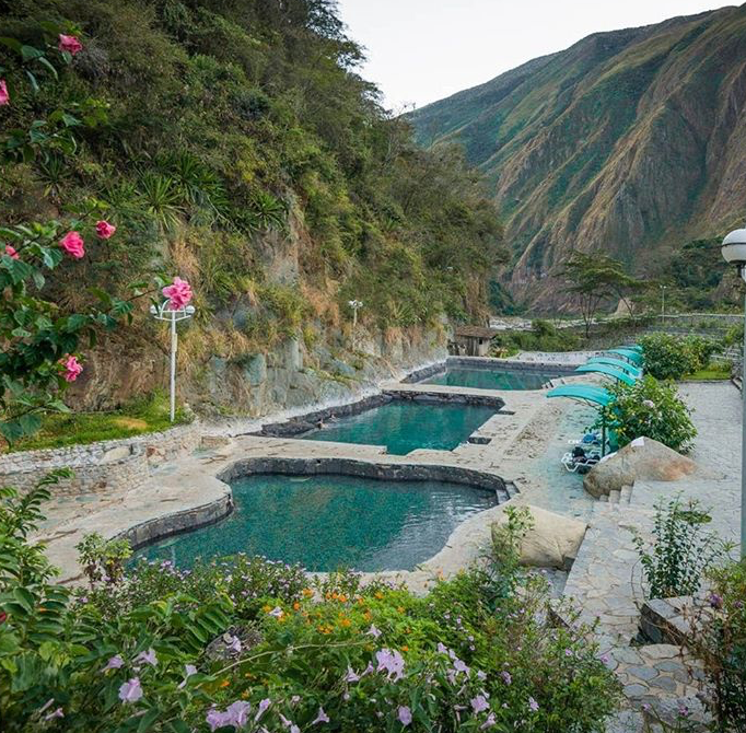 the image shows a series of outdoor hot springs pools nestled amidst lush vegetation and mountainous terrain. the pools are constructed with stone borders and filled with clear, turquoisecolored water. sun loungers and umbrellas are positioned near the pools, suggesting a relaxing spa or resort setting. the surrounding landscape is characterized by steep mountains covered in dense greenery and vibrant flowers. a stone path leads through the area, and a lamp post stands on the side, providing lighting.