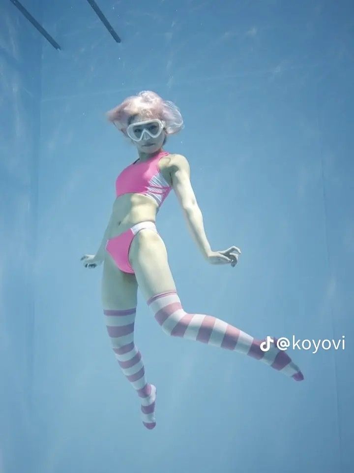 a woman in a pink and white cosplay swimsuit is posing underwater in a swimming pool. she's wearing a pink wig and white goggles. the background is a light blue.  the lighting is artificial and the perspective is from a low angle shot.