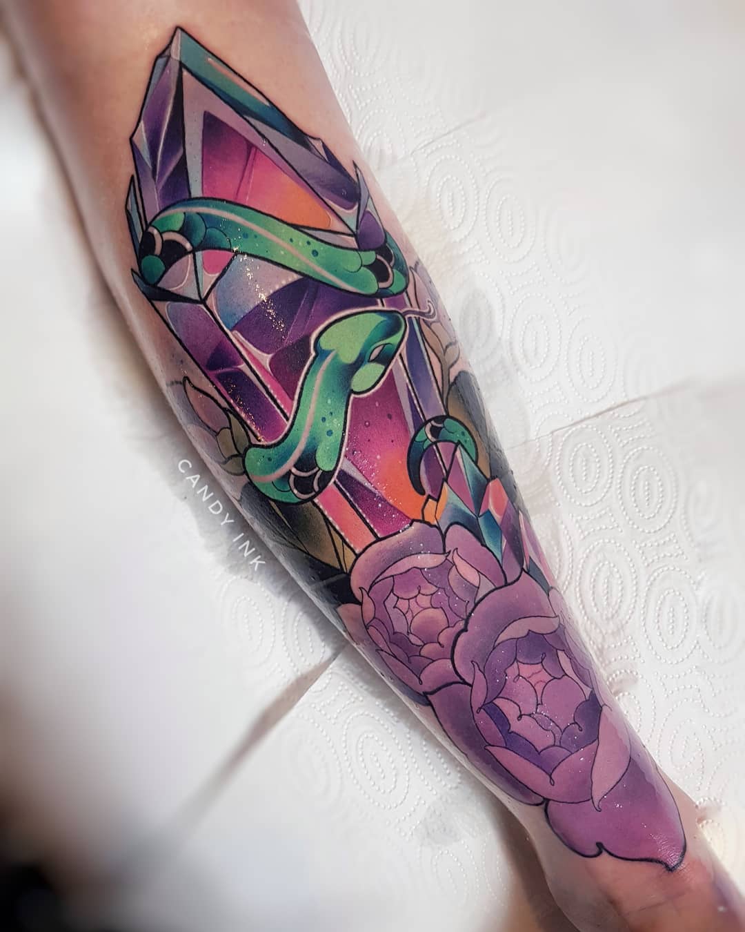 a colorful tattoo of a purple crystal with a snake wrapped around it and a purple rose at the bottom. the tattoo is on someone's lower leg.