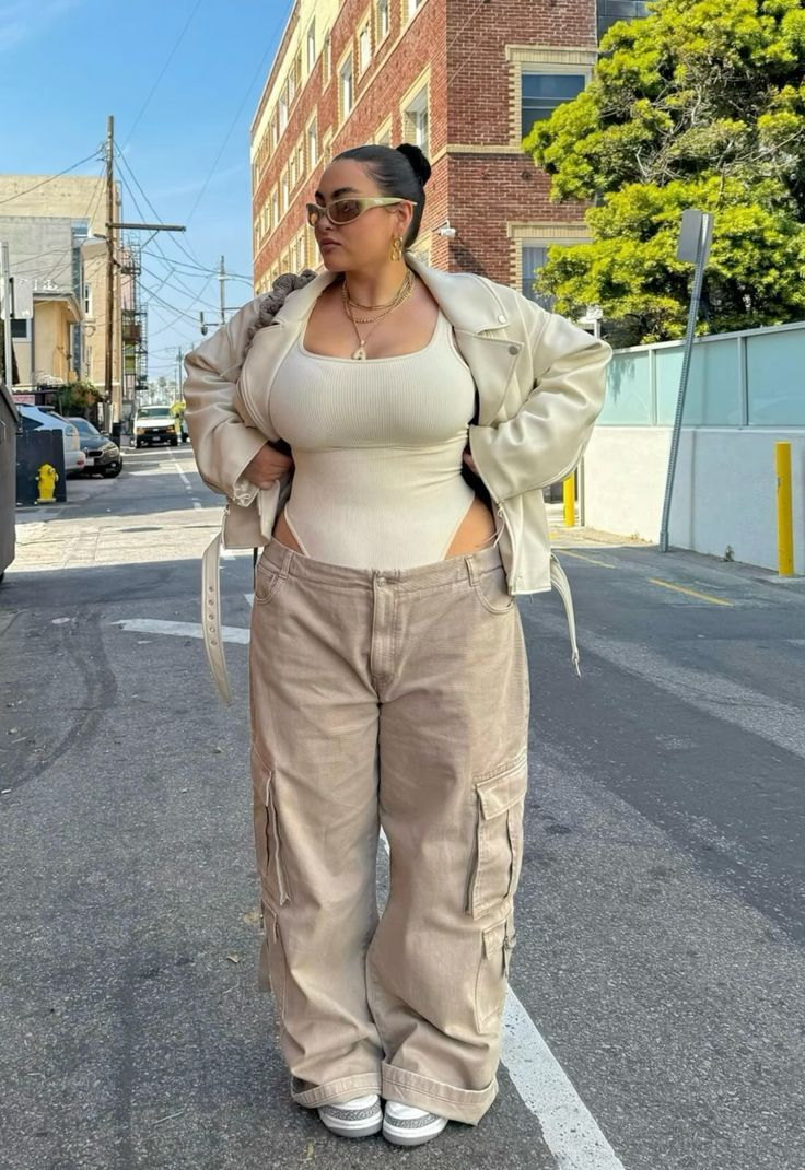 a fulllength portrait of a woman standing on a street. she is wearing a white bodysuit, beige cargo pants, a creamcolored jacket, sunglasses, and sneakers. she is wearing jewelry, including a necklace. the background shows buildings and a street scene.