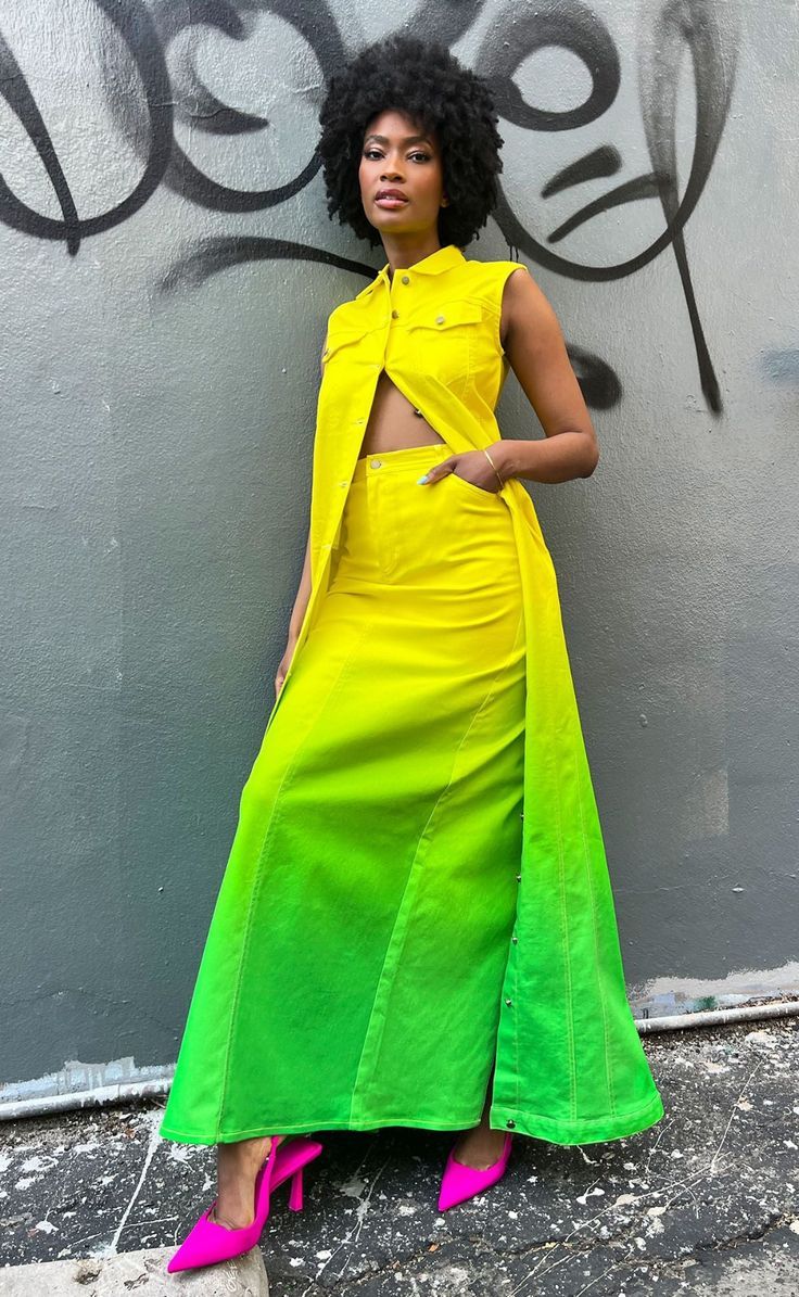 a stylish woman poses in a vibrant yellow and green ombre denim skirt and sleeveless shirt. she's wearing bright pink heels and standing against a wall with graffiti. she's  looking directly at the camera with a confident expression.