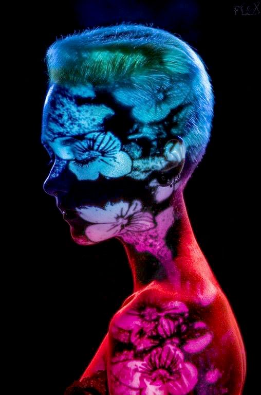 a closeup profile view of a woman with short, textured hair.  her body is adorned with intricate floral designs painted using uvreactive body paint. the colors are strikingly vibrant and neon, with blues, greens, and pinks dominating the image. the background is pure black, allowing the bright body paint to stand out dramatically. the overall style is artistic and somewhat surreal, showcasing the beauty of body art under blacklight. the image has a high contrast, emphasizing the details of the paint and the model's features.