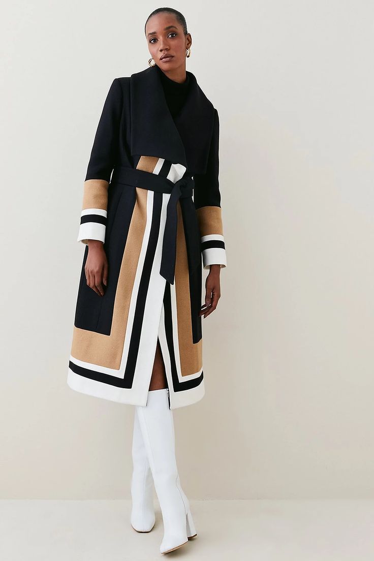 a woman in a black and white striped long coat with a brown belt, white kneehigh boots, and a black turtleneck stands in a studio, posing for the camera. she has dark hair, dark eyes, and a stylish, elegant look.