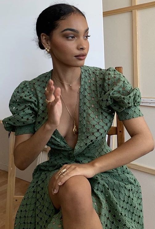 a woman sits in a chair wearing a green eyelet dress with puff sleeves. she is also wearing gold jewelry, including a necklace, earrings, and rings.