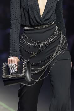 a woman is standing on a runway wearing a black dress with multiple chain belts and a black clutch bag. she has a stylish, glamorous look.