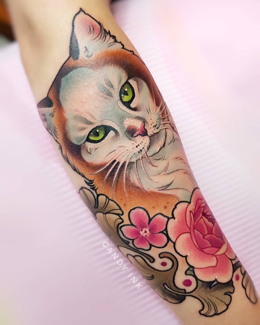 a neo traditional style tattoo of a cat with green eyes and flowers on someone's arm. the tattoo has pink and orange tones.
