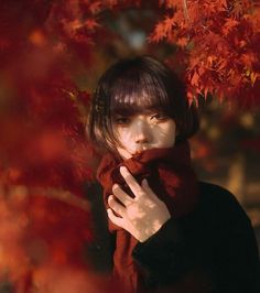 a young woman with dark hair and a red scarf is standing in front of a red leaf background. she is looking off to the side and has a sad, thoughtful expression on her face. the image is moody and atmospheric.