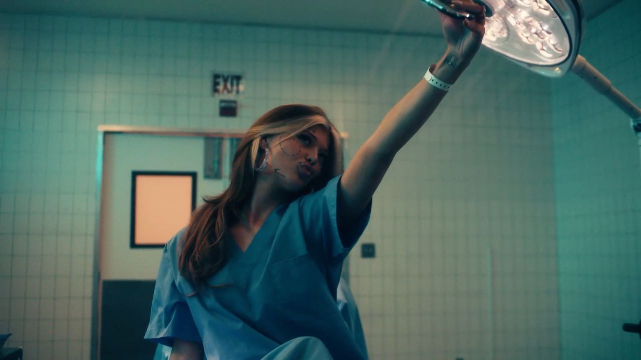 a woman in blue scrubs is taking a selfie in what appears to be a hospital operating room. she has blonde hair with a streak, and there are lines drawn on her face, likely indicating areas for cosmetic surgery. she's pouting and holding up her arm to take the photo, with a surgical lamp visible above. she's wearing a hospital bracelet. behind her, there is a tiled wall, an exit sign, and a door.