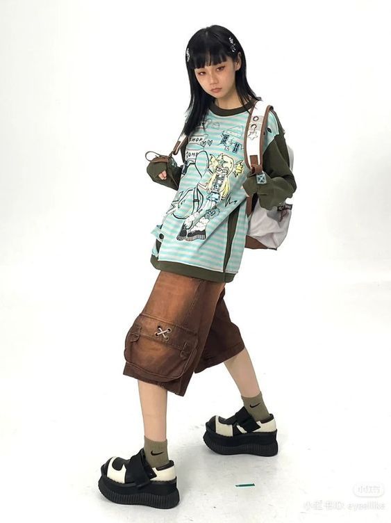 young asian girl is posing in a stylish outfit. she's wearing a light green longsleeved shirt with an anime graphic print, brown cargo shorts, and black platform shoes. she also has a brown and white backpack and is wearing her hair down. the background is plain white, which emphasizes the girl's outfit.
