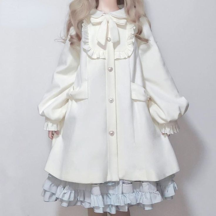 a person is wearing a white lolita fashion coat. the coat is long and has a ruffled collar with a bow. the coat also has large buttons and puffy sleeves. the person is also wearing a blue and white dress underneath the coat.  the image is taken indoors and the lighting is bright and soft.