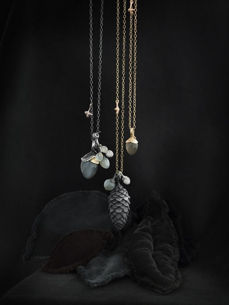 the image shows three necklaces with pendants against a dark background. the necklaces are made of silver and gold chains. the pendants are shaped like an acorn and a pine cone. the acorn pendant is made of silver and the pine cone pendant is made of gold. the necklaces are hanging above some black cloth shaped like leaves.