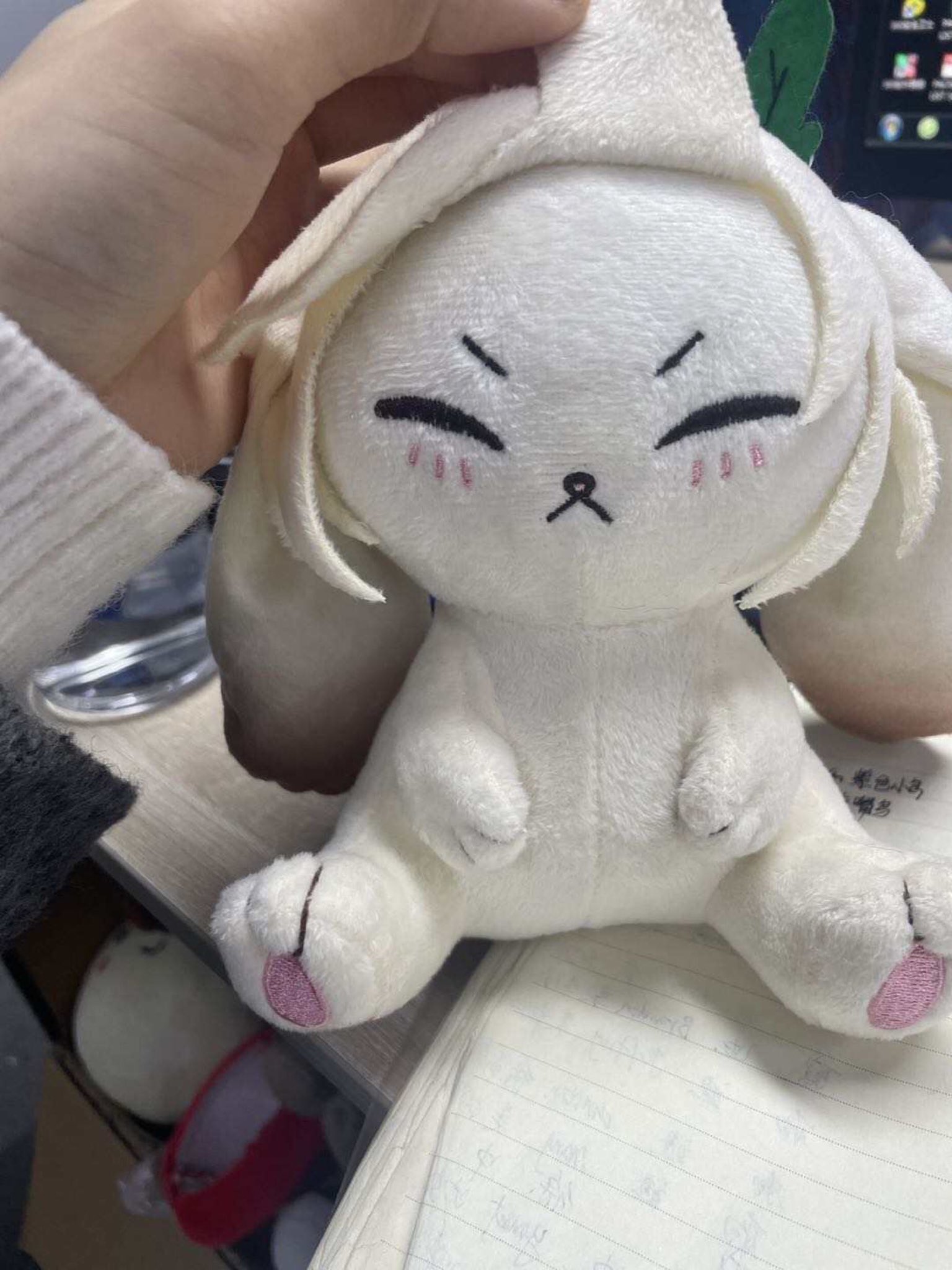 a closeup of a white plush bunny with large, cartoon eyes and pink cheeks. the bunny is sitting on a notebook with its head tilted to the side. it is made of soft, fluffy material and has a sweet, innocent expression.