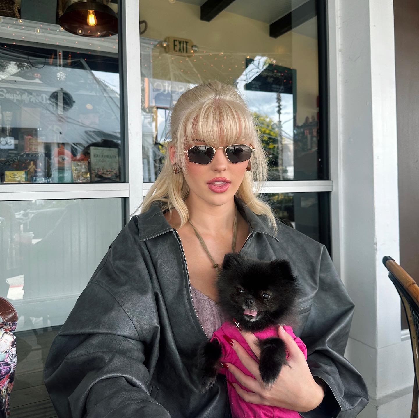 a woman with blonde hair and bangs is sitting at a table, holding a small black pomeranian dog. the woman is wearing sunglasses, a gray jacket, a necklace, and earrings. the dog is wearing a pink outfit. they are at a restaurant or cafe, as there is an "exit" sign in the window behind them and a table.