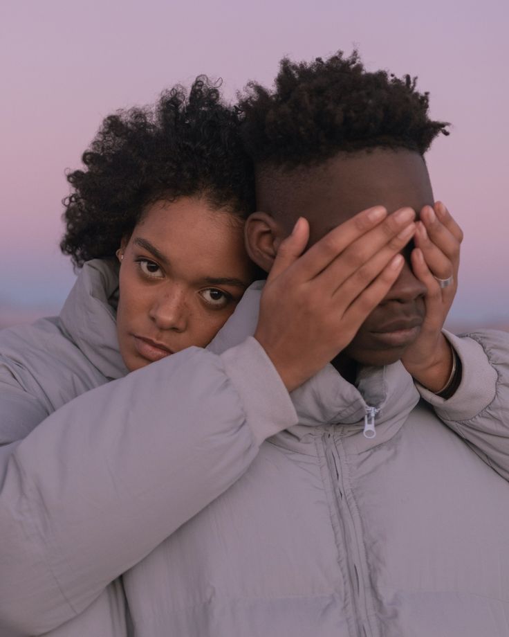 a young black couple is posing for a portrait. the man is wearing a grey jacket and has his hand over his eyes. the woman is wearing a grey jacket and has her arm around the man. they are both looking at the camera and smiling. they appear to be deeply in love and happy together.