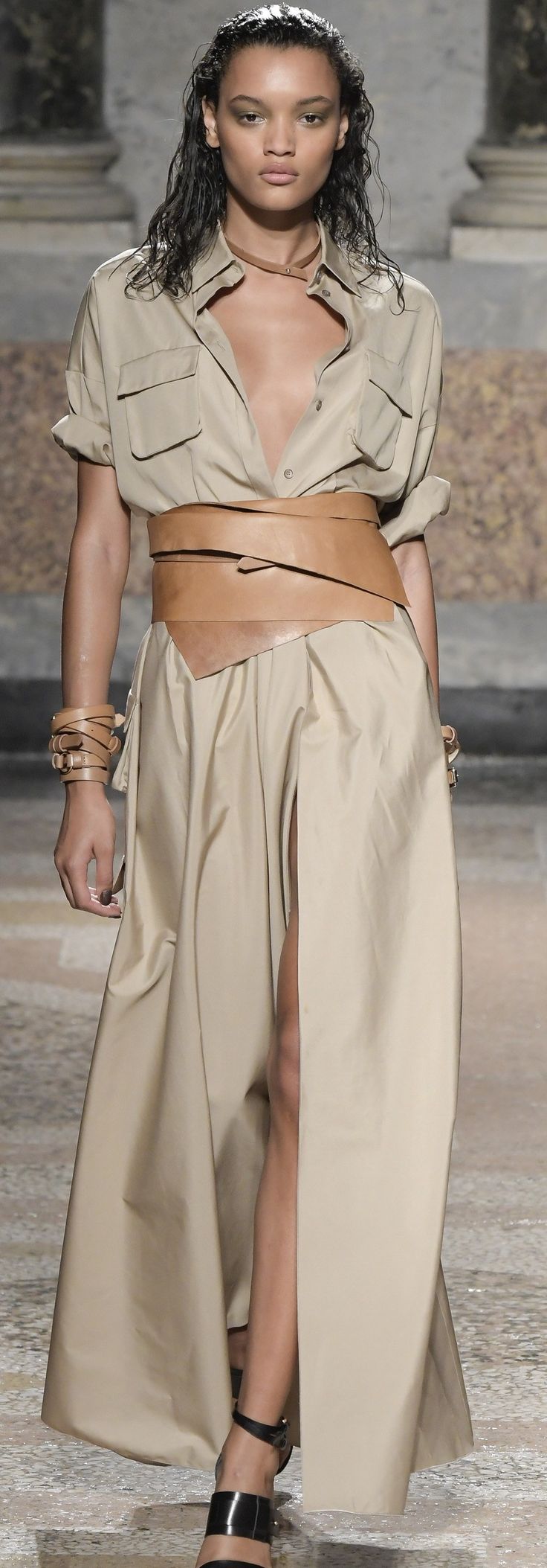 a full shot of a female model showcasing a beige long shirt dress on a runway. the dress features a high waistline accentuated by a wide brown leather belt wrapped around it. the model has long dark hair styled loosely, and she's wearing simple black sandals. the setting appears to be an outdoor runway, with stone structures visible in the background. the overall style of the dress and the model's appearance are elegant and sophisticated.