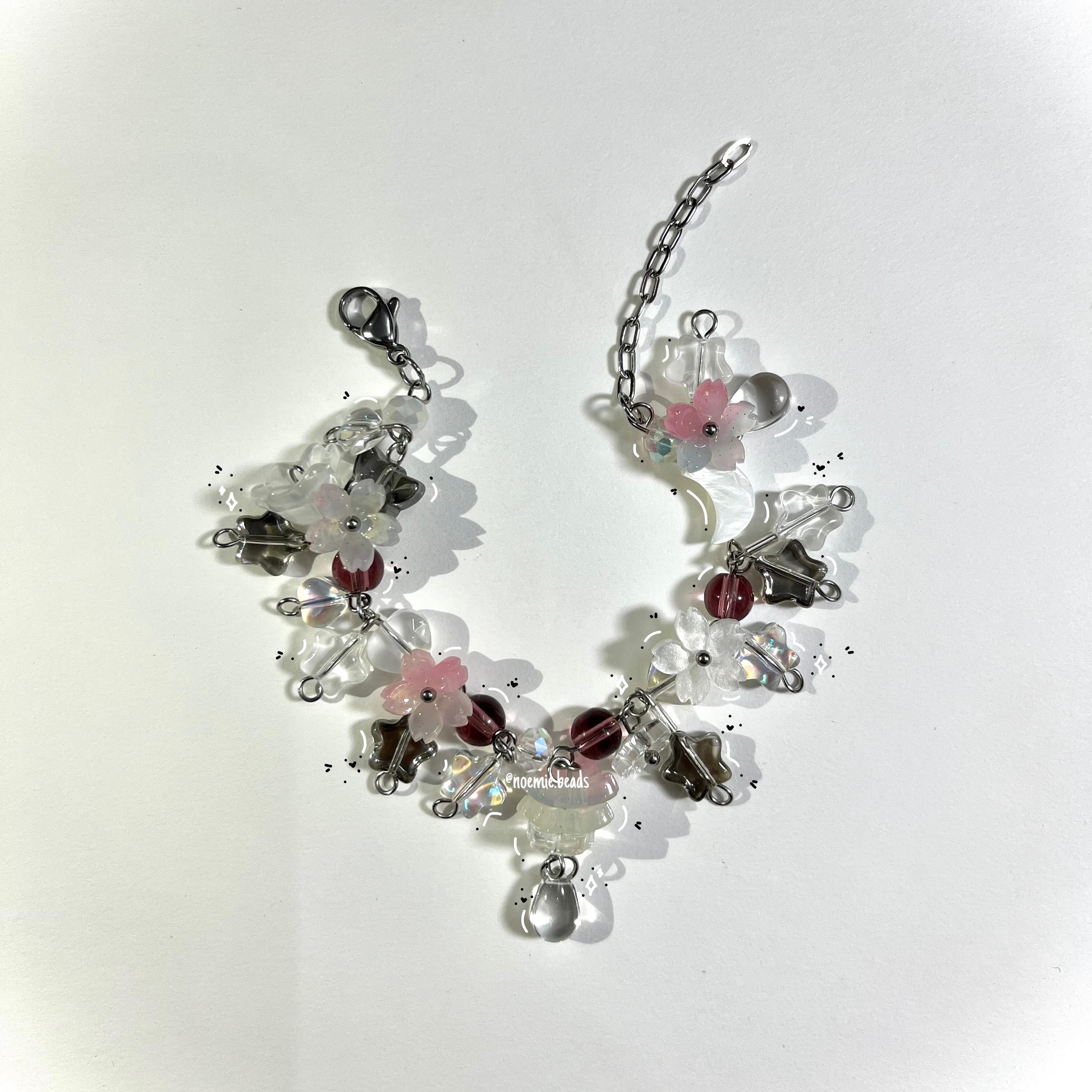 a handmade beaded bracelet featuring a variety of beads, including clear, white, pink, and gray, along with flowershaped beads and a silver chain with a lobster clasp closure, casting shadows on a white surface.