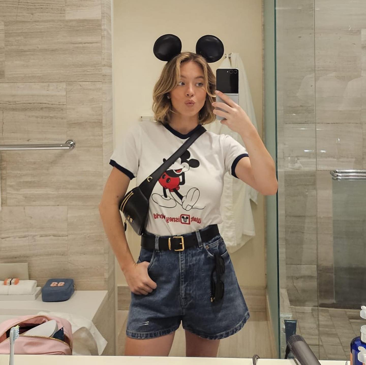 a young woman takes a selfie in a bathroom mirror. she is wearing a white tshirt with a mickey mouse graphic, denim shorts, a black crossbody bag, and mickey mouse ears. she's smiling and appears happy. the setting appears to be a hotel bathroom.  she is holding a cell phone in her hand.