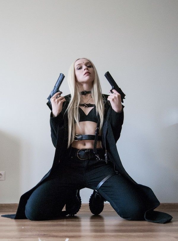 a blonde woman is kneeling on the floor in a dark, allblack outfit, holding two black pistols in her hands. she is looking directly at the camera with a serious expression. she's wearing a long black coat, a black leather harness, and black pants.