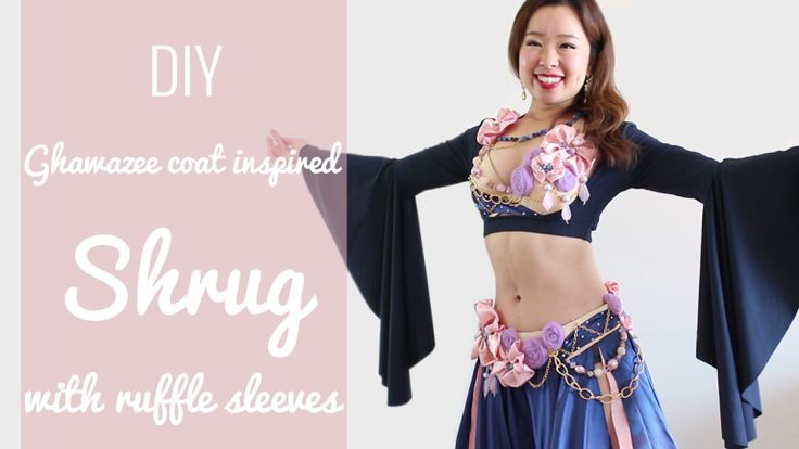a woman in a belly dance costume smiles broadly, standing against a white backdrop. she is wearing a dark blue crop top with long, fluted sleeves, a matching skirt with pink accents, and a heavily embellished bra and belt adorned with pink and purple flowers. text overlay on the left side of the image reads "diy ghawazee coat inspired shrug with ruffle sleeves" in a decorative, light pink font, suggesting this is a presentation or tutorial for creating the costume.