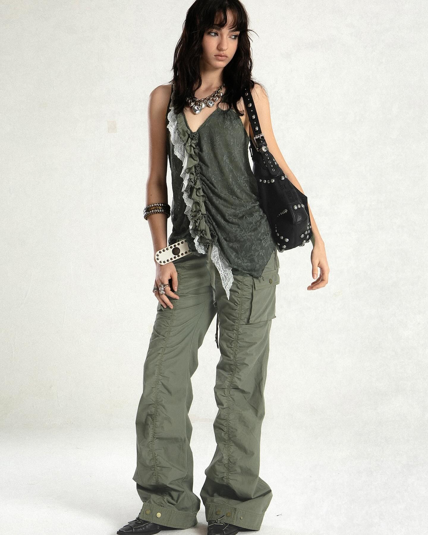 a fulllength shot of a young woman, standing against a white background, showcases a stylish bohemiangrunge outfit. she wears olive green cargo pants, a greyishgreen ruffled top with lace detailing, several silver accessories including a necklace and bracelets, and a studded black shoulder bag. her long dark brown hair is loose around her shoulders.  the overall style is casual yet chic and slightly edgy. the model is posing in a relaxed manner, and her expression is natural and slightly introspective. the lighting is even, enhancing the details of her clothing and accessories.