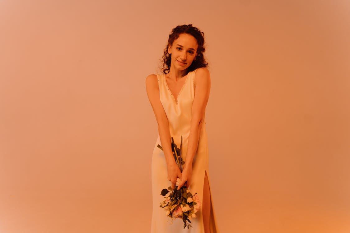 a young woman, possibly a bride, stands against a soft orange background. she's wearing a simple, elegant, offwhite sleeveless wedding dress with a thighhigh slit on one side. her curly brown hair is styled naturally, and she holds a small bouquet of lightcolored flowers close to her body. she looks directly at the camera with a gentle, happy smile. the overall mood is serene and romantic.  the lighting is soft and even, illuminating her features and the dress beautifully.