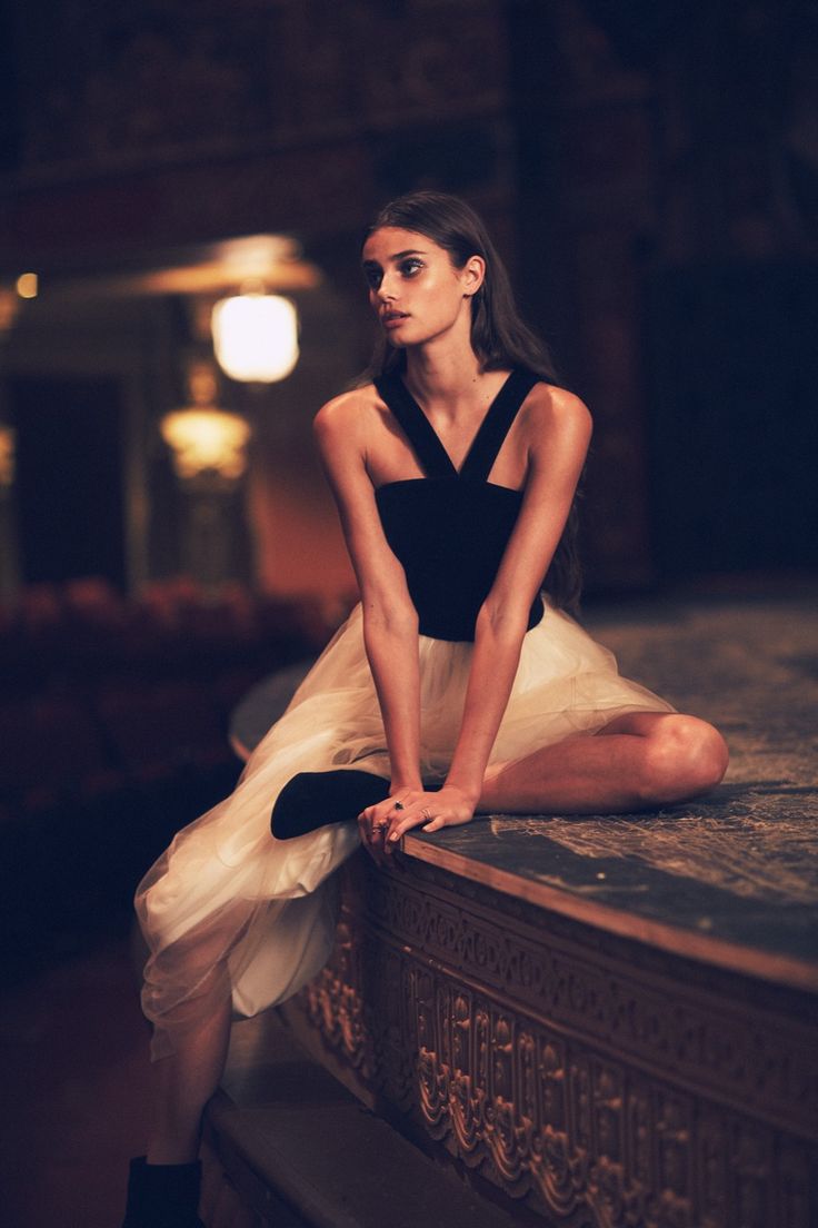 a woman is sitting on a stage in a dark theater. she is wearing a white tulle skirt and a black top. she is looking away from the camera, her arms are resting on her legs. she is wearing black ankle boots and has a ring on her finger. the lighting is dramatic and the background is dark, but the woman is welllit and her pose is elegant.