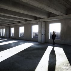 a large, open, and empty industrial room with concrete walls and floors. sunlight streams in through large windows, casting long shadows on the floor. a lone figure stands in the center of the room, silhouetted against the light.