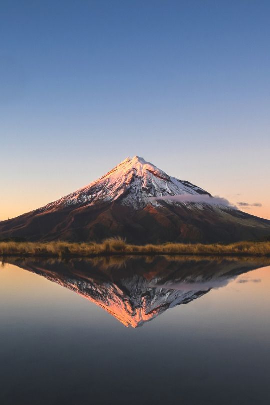 a snowcapped mountain with its reflection in a calm lake. the sky is a clear blue and the sun is setting behind the mountain. the mountain is in the center of the image and the lake is in the foreground.  the image is taken from a low angle and the mountain appears to be very large and imposing.