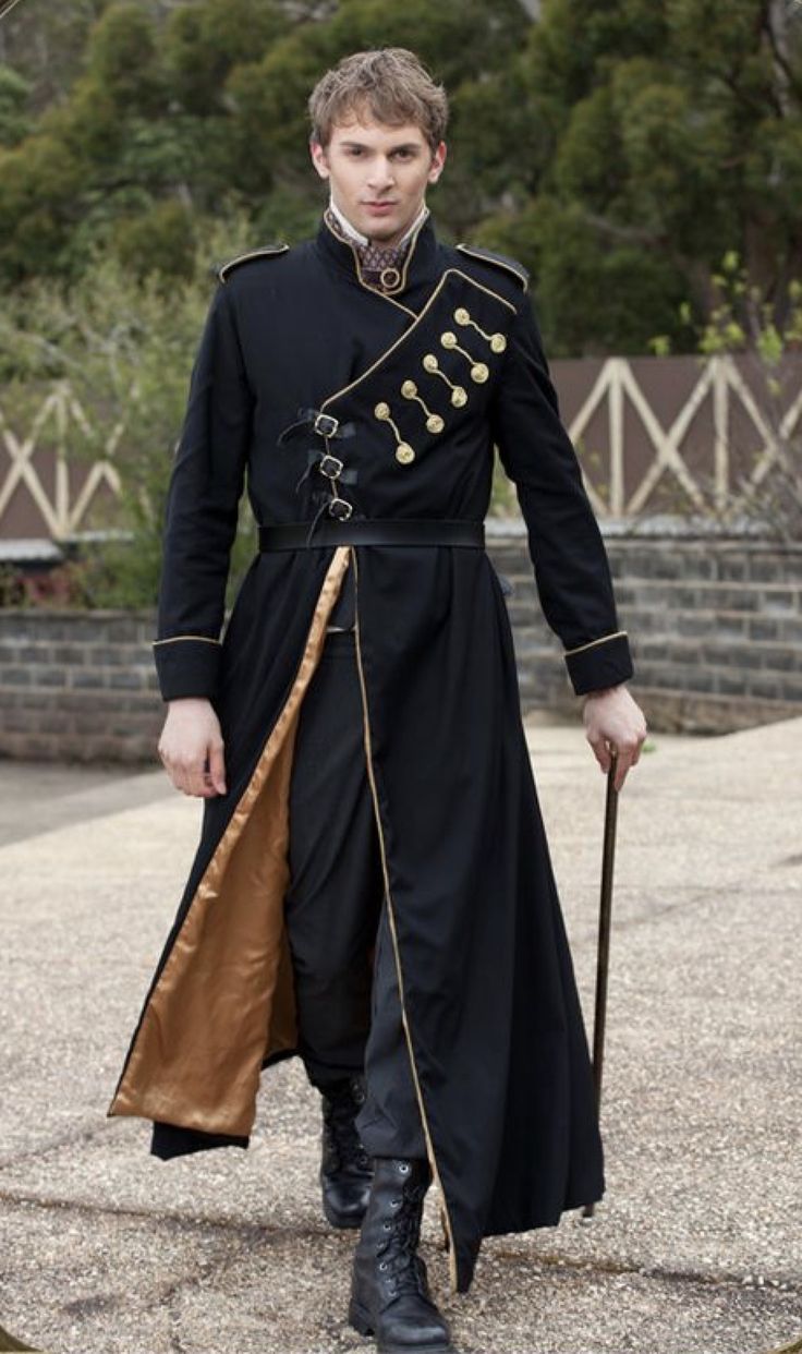 a man is standing outdoors wearing a black steampunkstyle coat with gold trim and gold buttons, and black boots. he is holding a stick in his right hand.