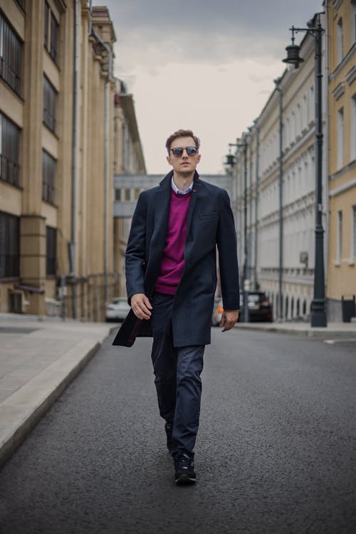 a stylish man in a dark overcoat, sunglasses, and a magenta sweater walks down a city street. the background features tall buildings and a cloudy sky. the overall mood is sophisticated and confident.