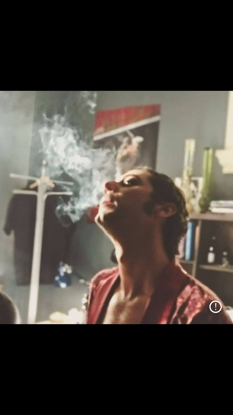 the image captures freddie mercury in a candid moment, likely backstage or in a private setting. he is shown in profile, head tilted back as he exhales a plume of smoke. he's wearing a patterned burgundy robe, contrasting against the muted colors of the cluttered room. the presence of a clothing rack and posters suggests a backstage area. the lighting is soft and natural, creating an intimate and somewhat nostalgic atmosphere.