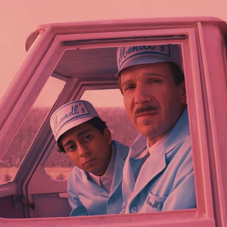 two men in white and blue ice cream truck uniforms and hats, looking at the camera. they are sitting in an ice cream truck. the image is taken through the window of the truck.