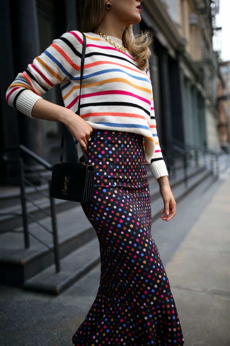 a stylish woman is standing on a city street wearing a colorful outfit. she is wearing a horizontal striped sweater in white, black, orange, pink, and blue. the sweater is paired with a black mermaid skirt covered in colorful polka dots. she carries a black yves saint laurent crossbody bag and wears gold necklaces and earrings. the background features a grey building with a black staircase. the photo is taken from a slightly low angle.