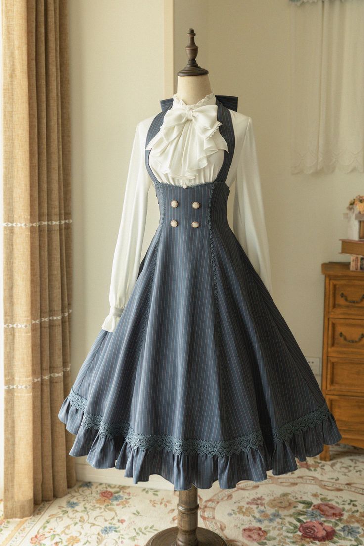 a navy blue pinstriped lolita fashion dress is displayed on a mannequin. the dress features a white blouse with a bow tie underneath, a high waist with decorative buttons, and a flowing skirt with lace trim. the mannequin is positioned in a welllit room with a window and a wooden cabinet in the background.
