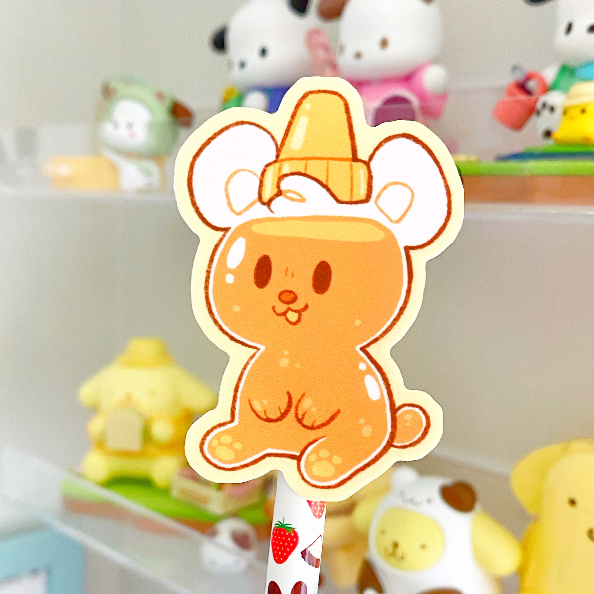 the image shows a closeup of a cute cartoon honey bear sticker affixed to a pencil. the bear is depicted as a small, round character with orange honeylike coloring and a small yellow honey pot on its head. the background is blurred but shows a collection of other cute plushies and stationery items, suggesting a shelf or desk organization system. the overall style is kawaii, characterized by its soft colors, rounded shapes, and adorable aesthetic. the sticker appears to be a collectible item.