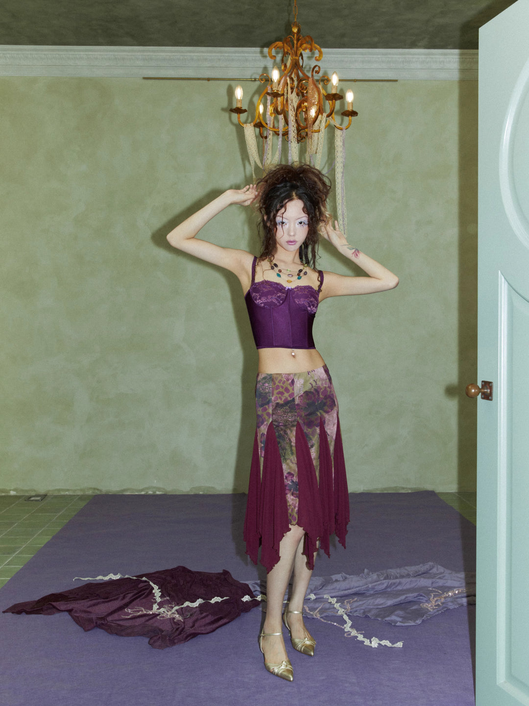 a young woman with dark hair is standing in a room with green walls. she is wearing a purple corset top and a patterned skirt with a burgundy overlay.  she is also wearing gold heels and multiple necklaces. an antique chandelier hangs above her. the floor is purple. there are pieces of fabric on the floor around her. the overall style is vintage or retro.